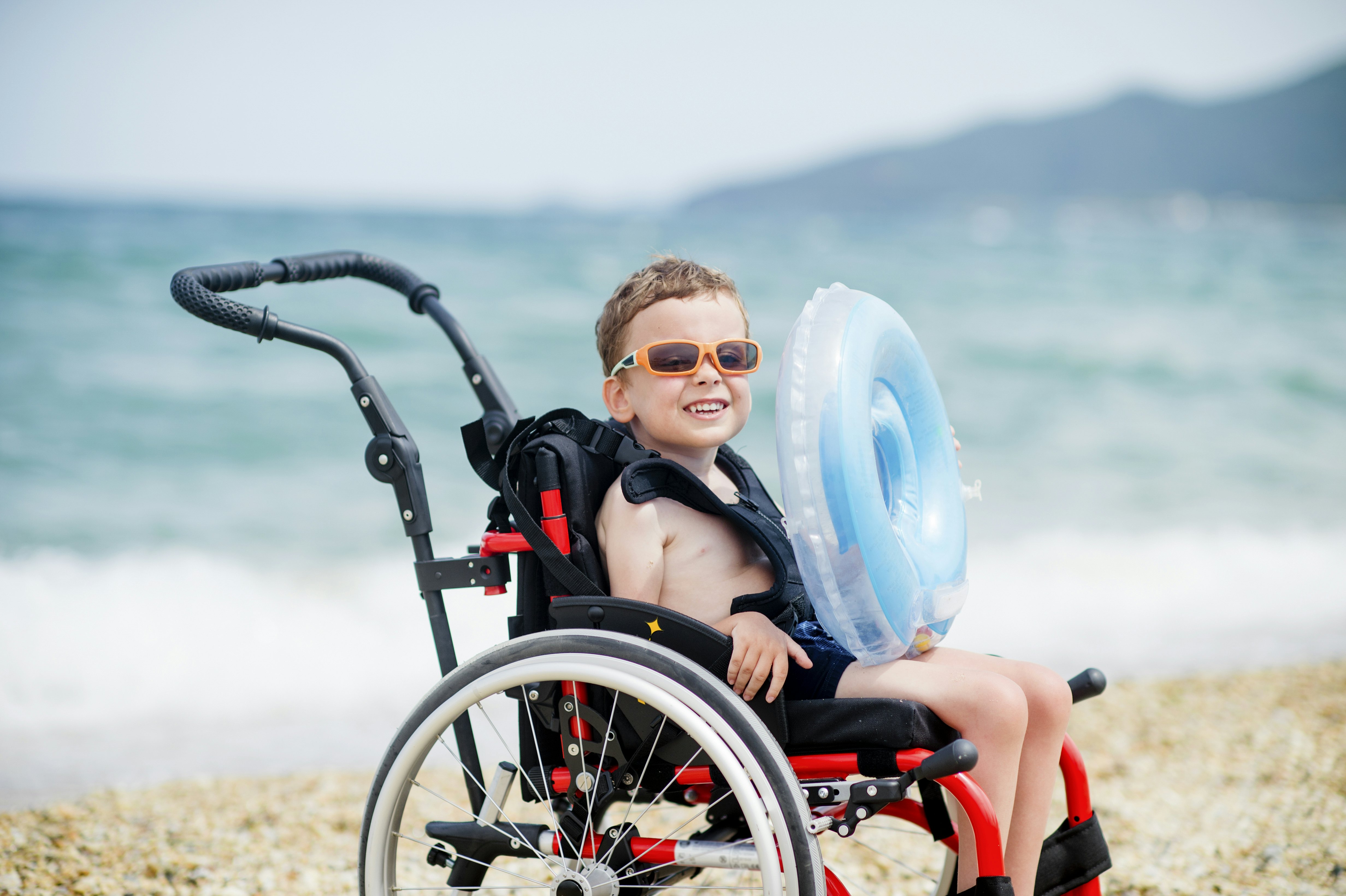 A cute boy holding an inflatable floaty in wheelchair going to swim.