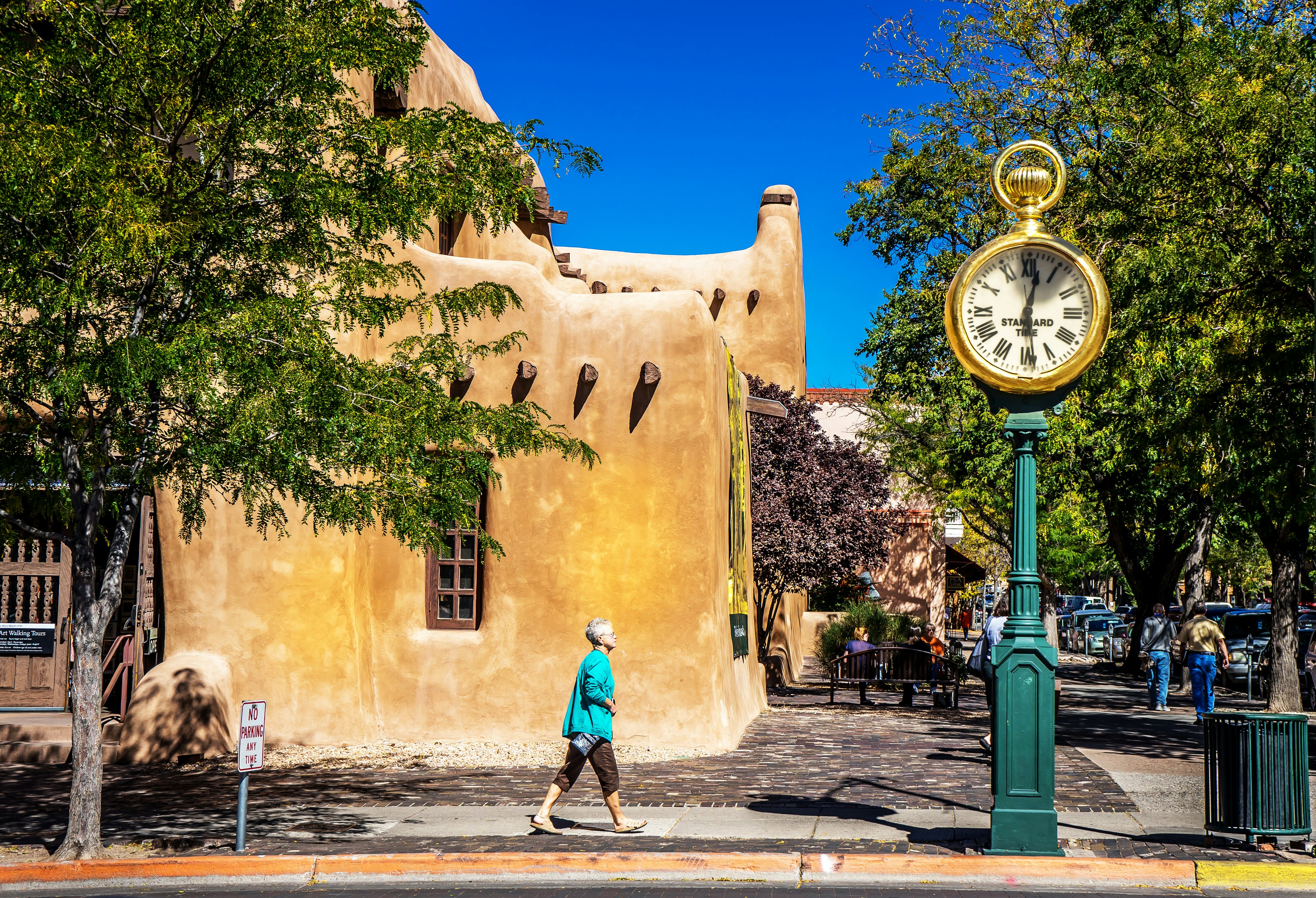 A woman in a turquoise tunic walks in front of an orange adobe building in a city street. A large clock is on the street corner.