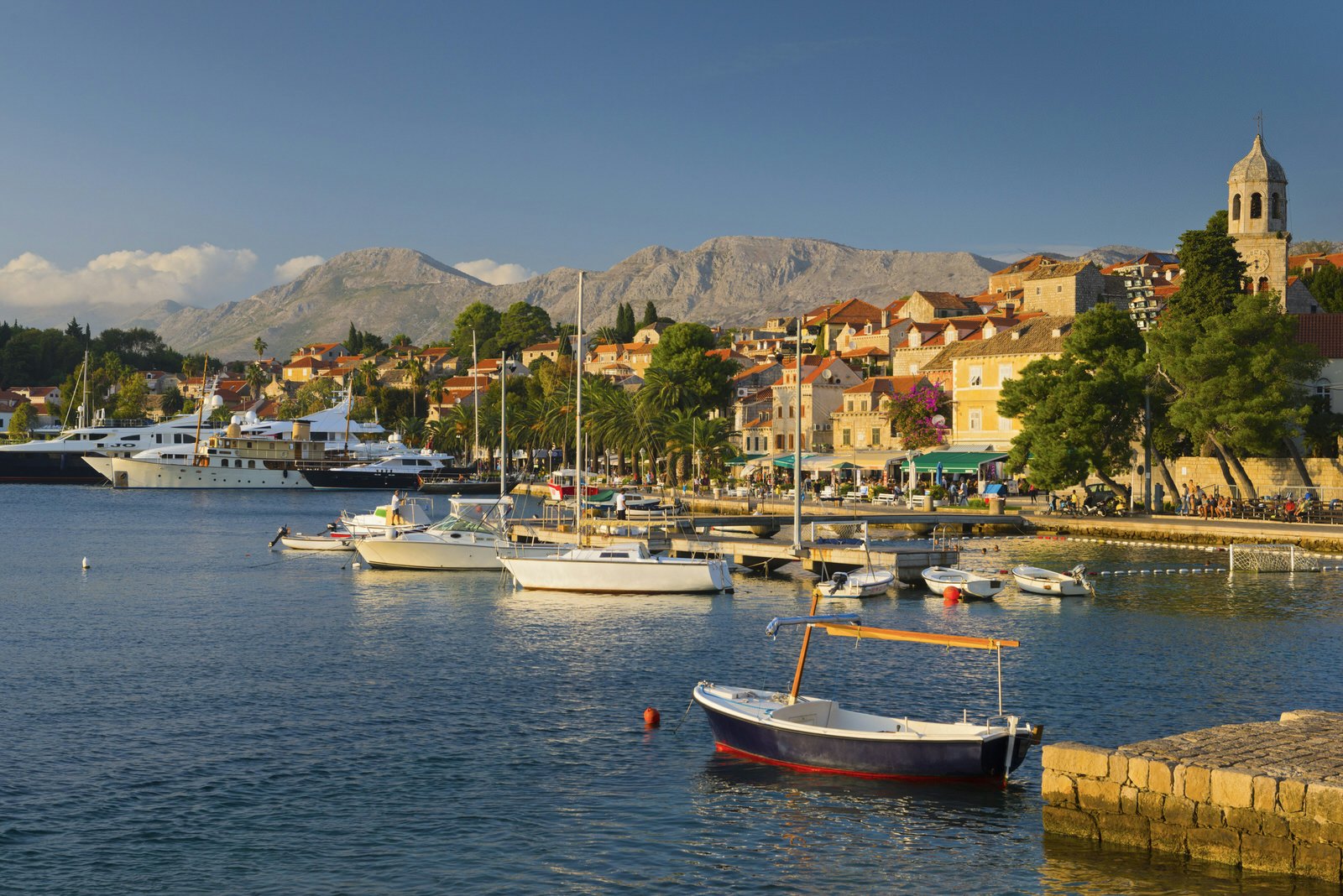 Cavtat's pretty marina under late afternoon sun: boats are moored on the water, with a stone church spire, orange-roofed buildings and trees along the shore; barren mountain peaks form a dramatic backdrop.