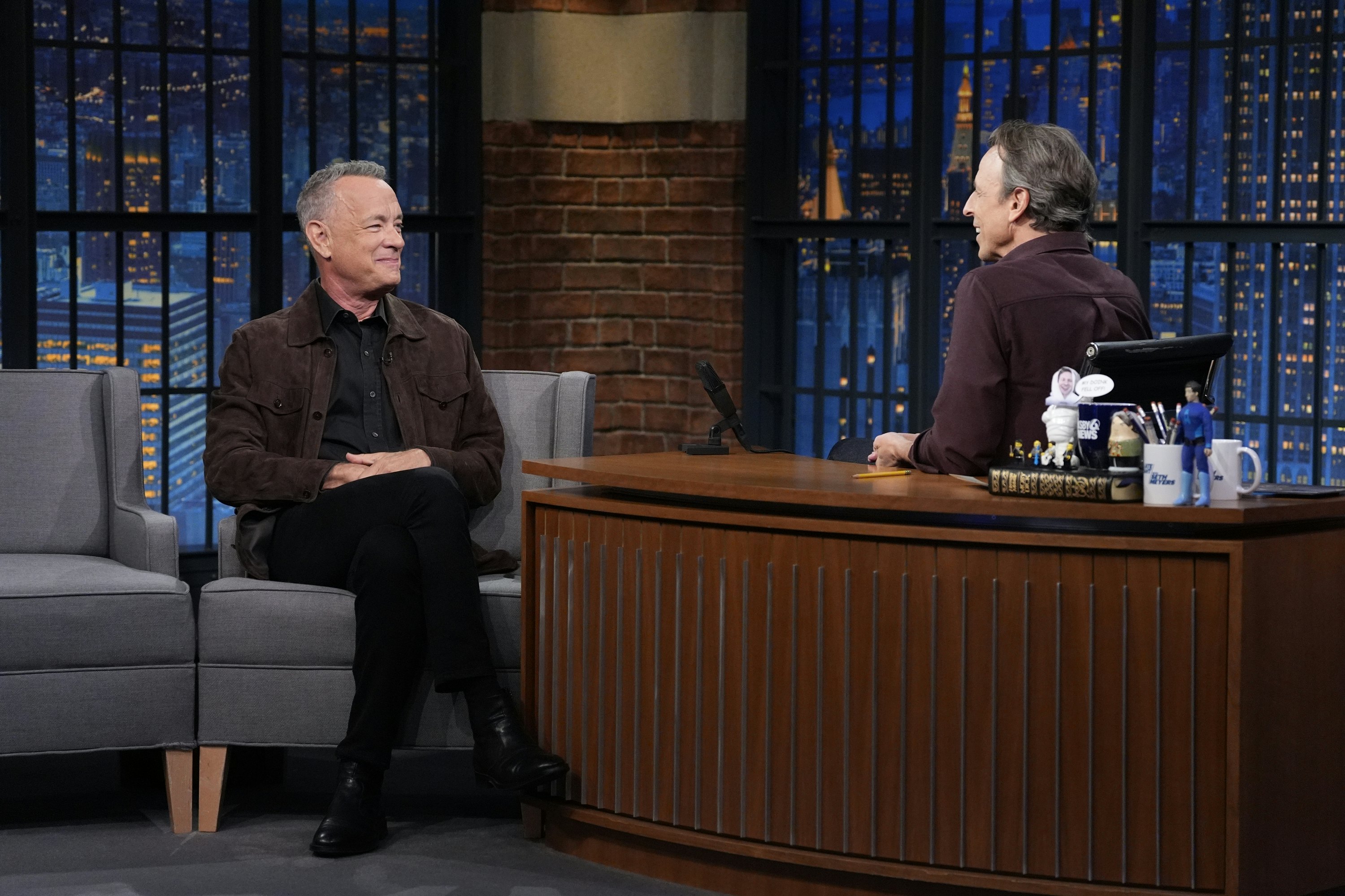 Two men sit in a TV studio in discussion. One is actor Tom Hanks, the other is the host, Seth Meyers