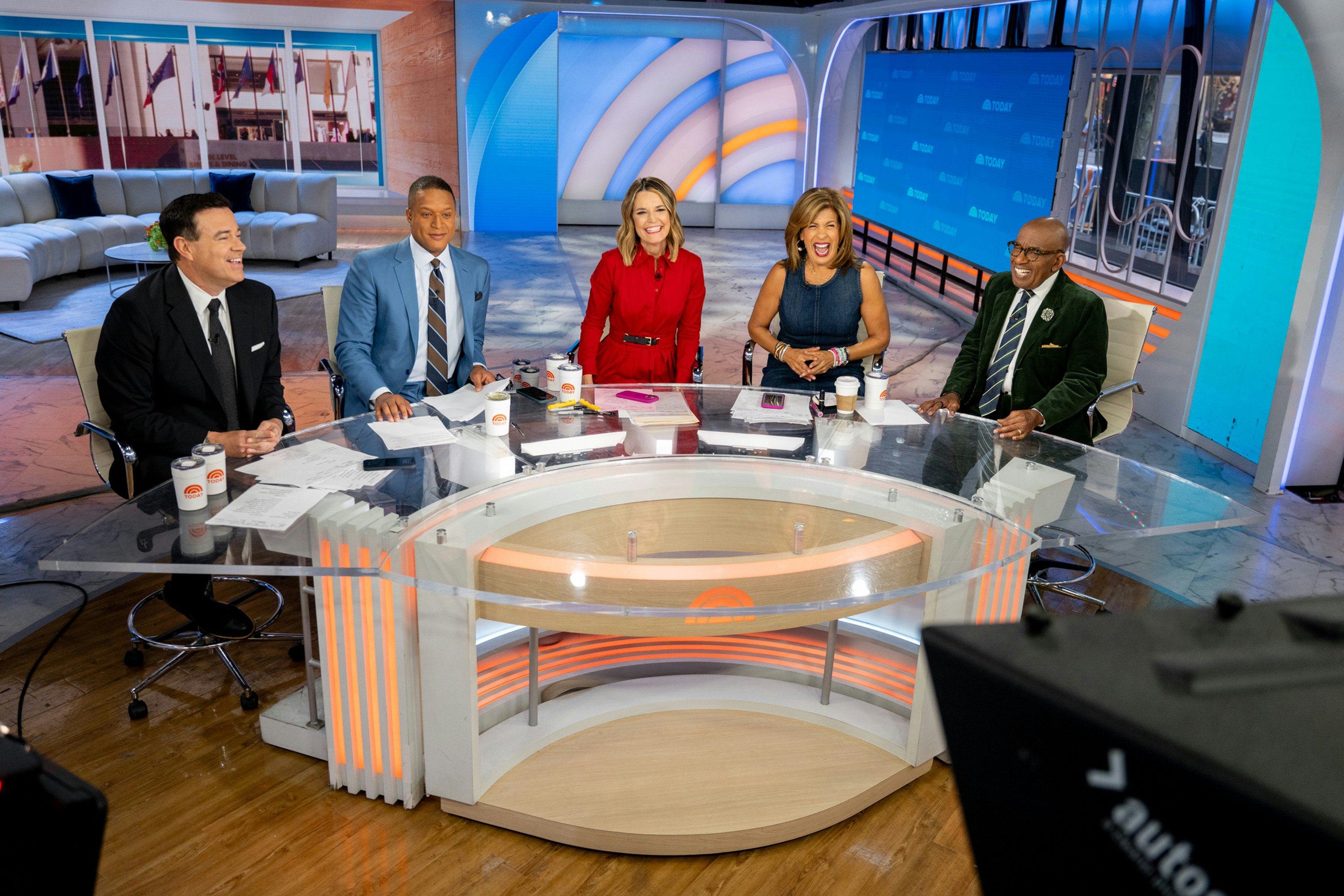 A panel of TV hosts smile as they sit around a large glass desk in a studio.