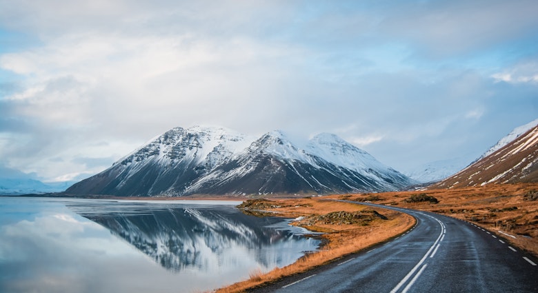Panoramic winter photo of road leading along coast of lake to volcanic mountains. High rocky peaks covered with snow layer mirroring on water surface. Driver's point of view on Ring road, Iceland. License Type: media Download Time: 2023-08-01T15:17:30.000Z User: FergalCo Is Editorial: No purchase_order: