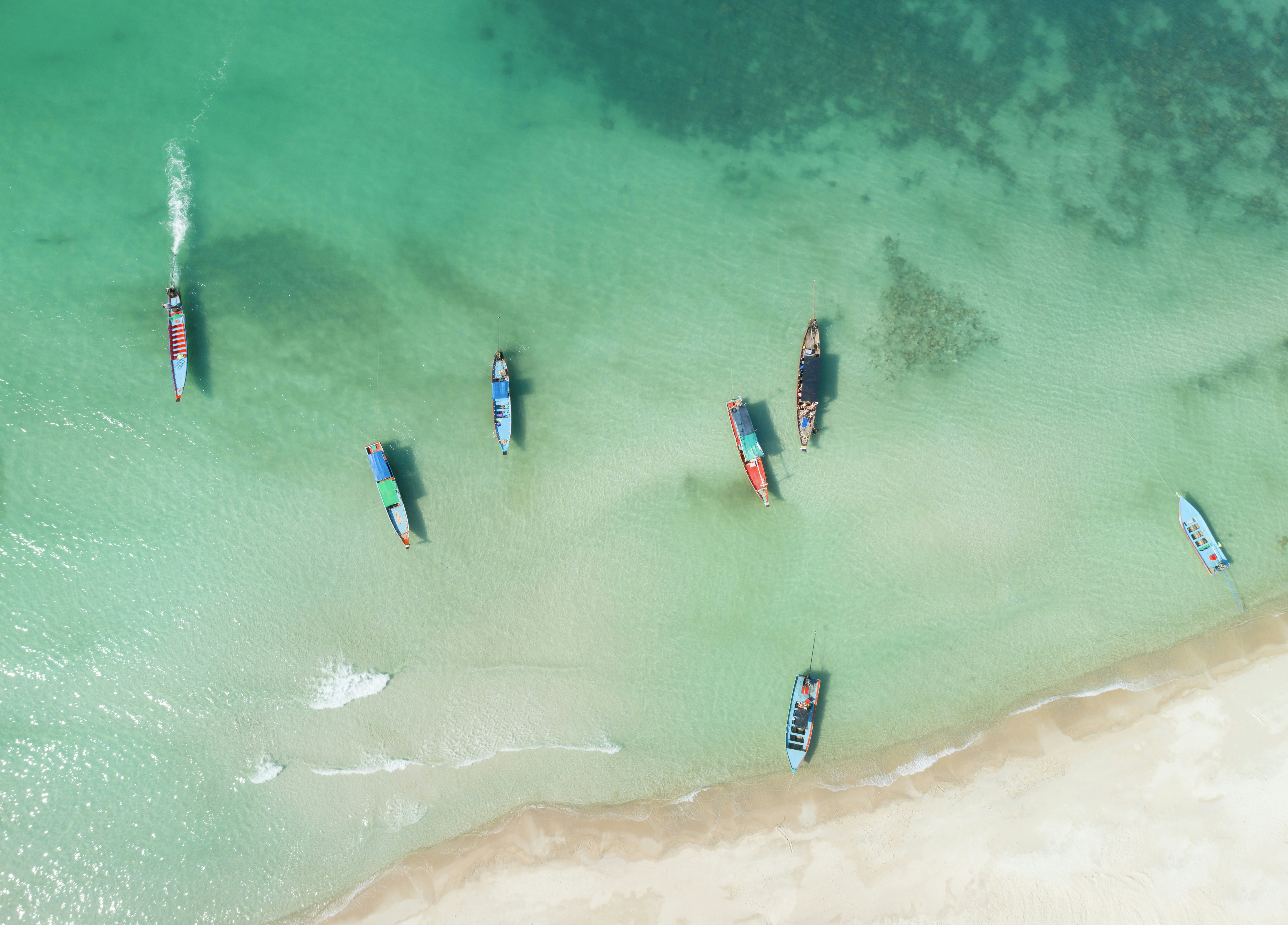 An aerial view of colorful fishing boats moored in shallow turquoise waters next to a white-sand beach