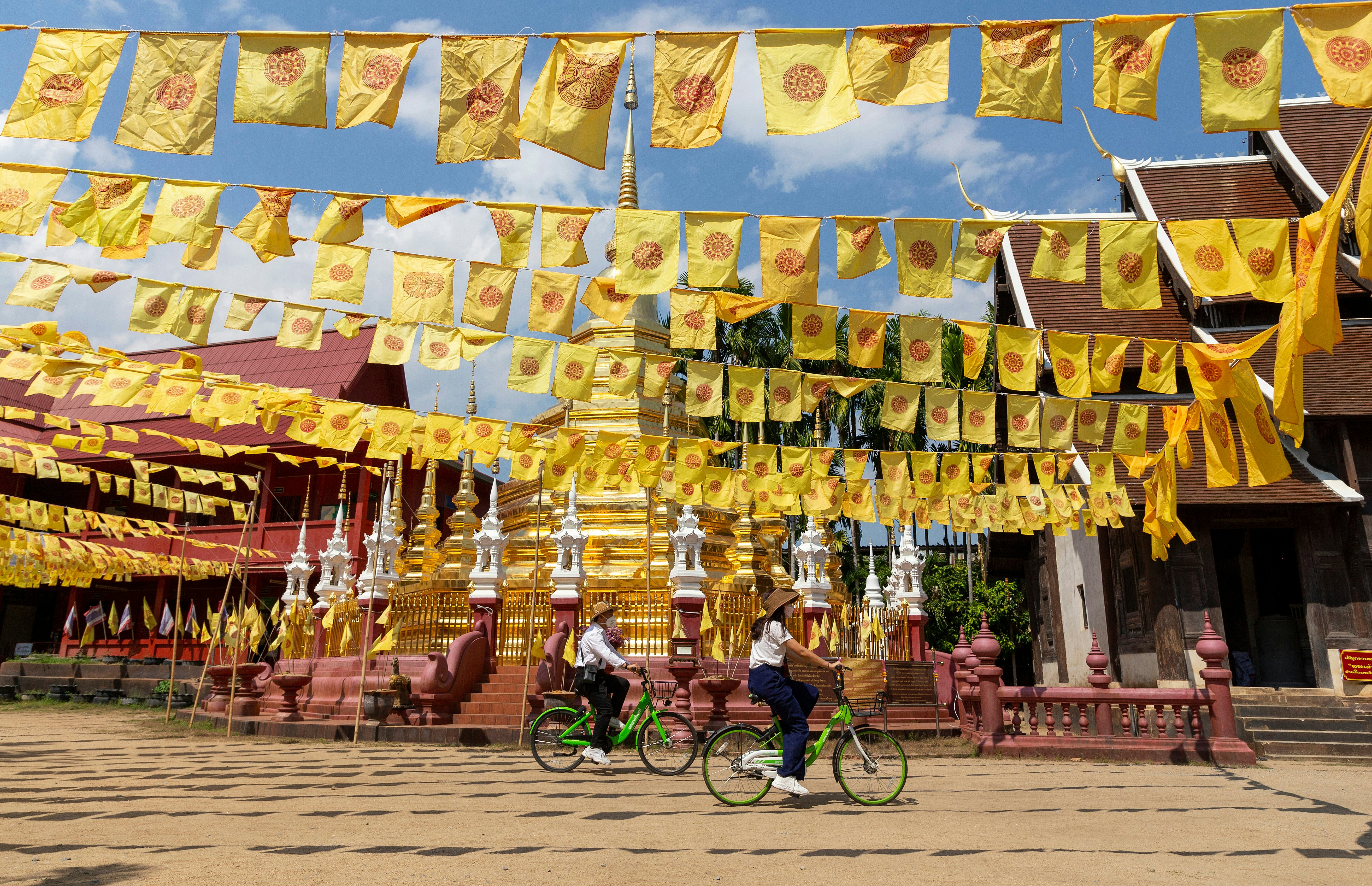 Two people on bicycles pedal past the gold stupa of a temple. Numerous yellow flags hanging from strings flutter overhead.