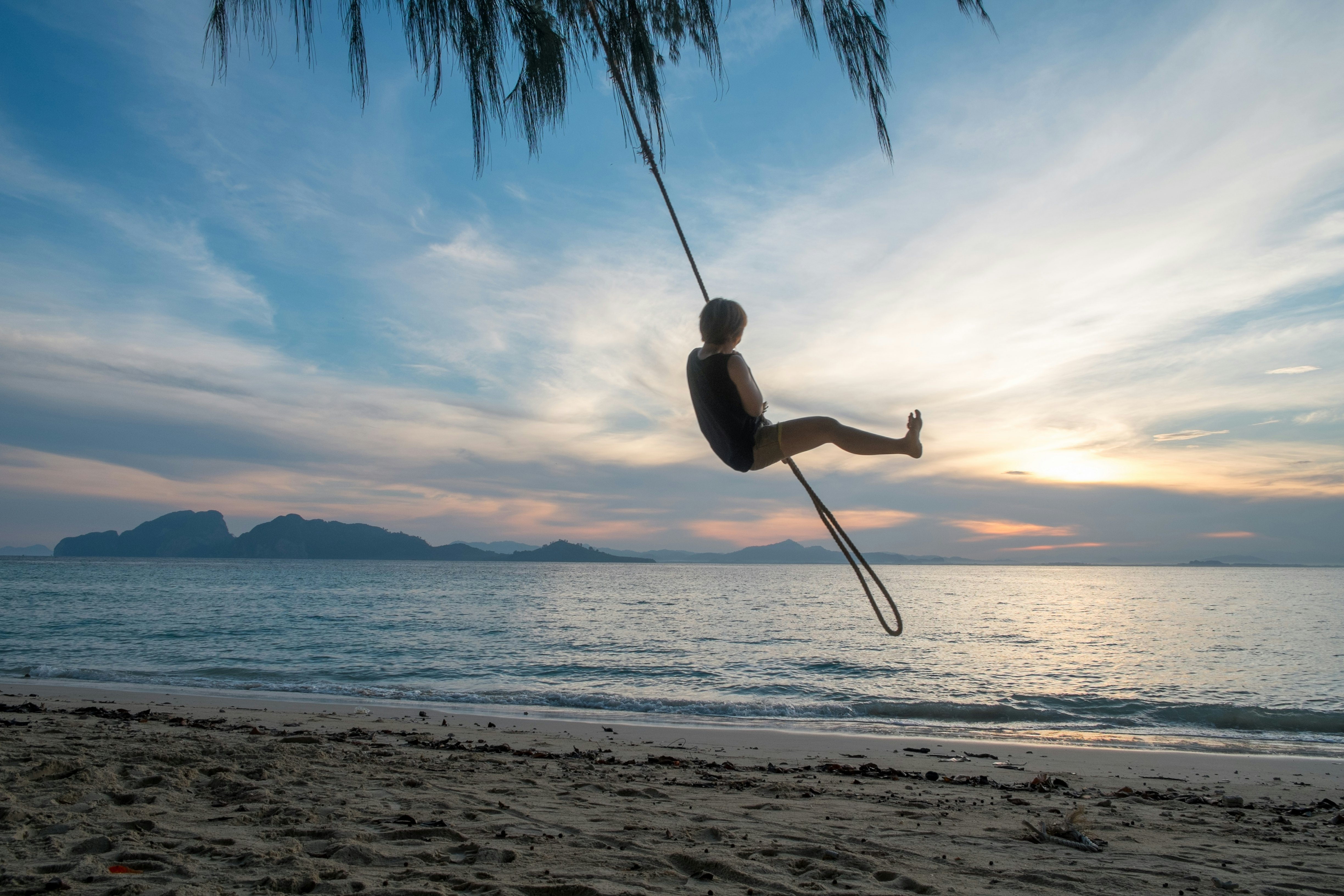 A young person hang from a rope swing above a beach, looking out at the post-sunset light over the water and islands beyond