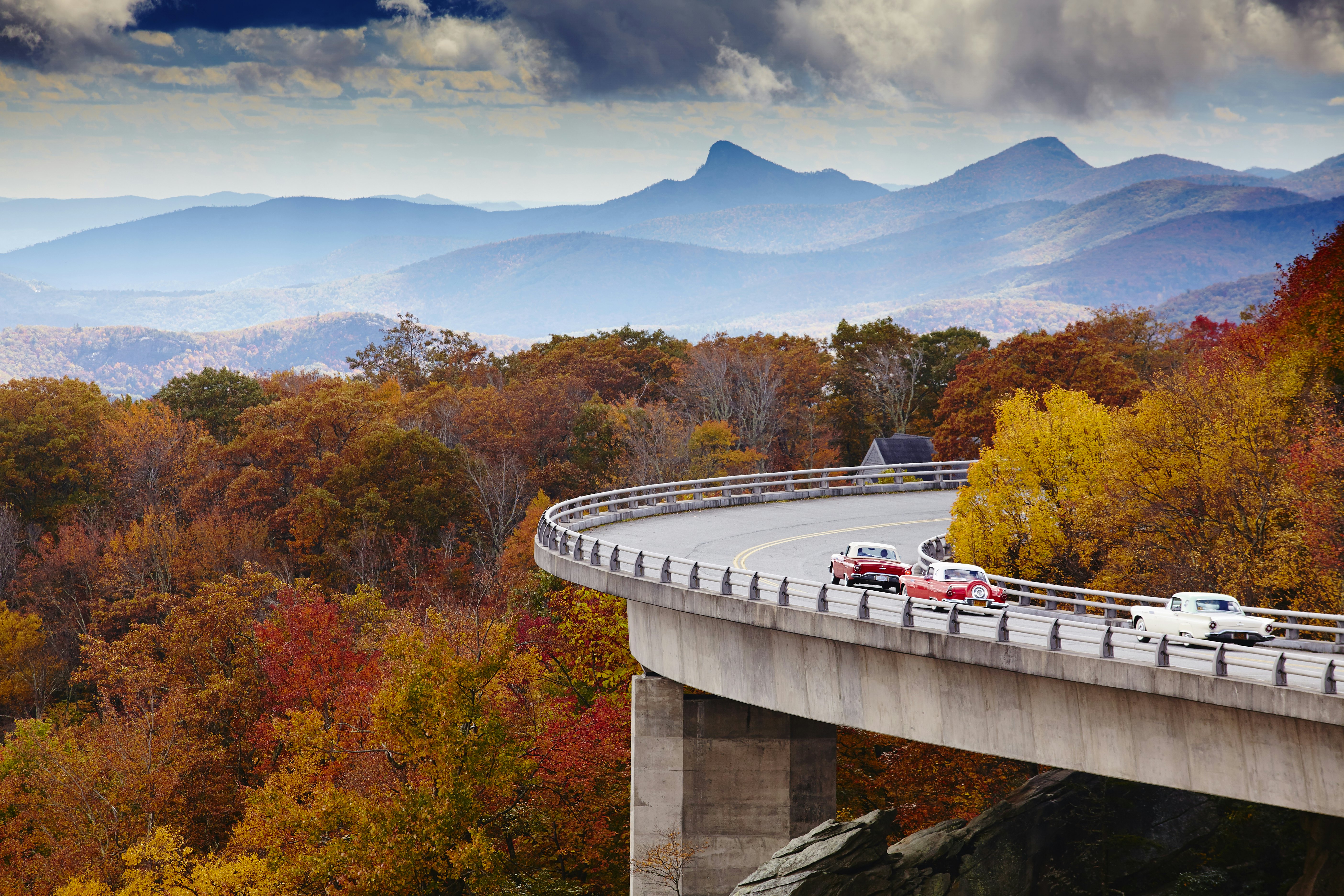 Vintage cars drive along a curved road viaduct on a hillside covered with trees showing fall foliage. Hazy mountains are visible in the distance.