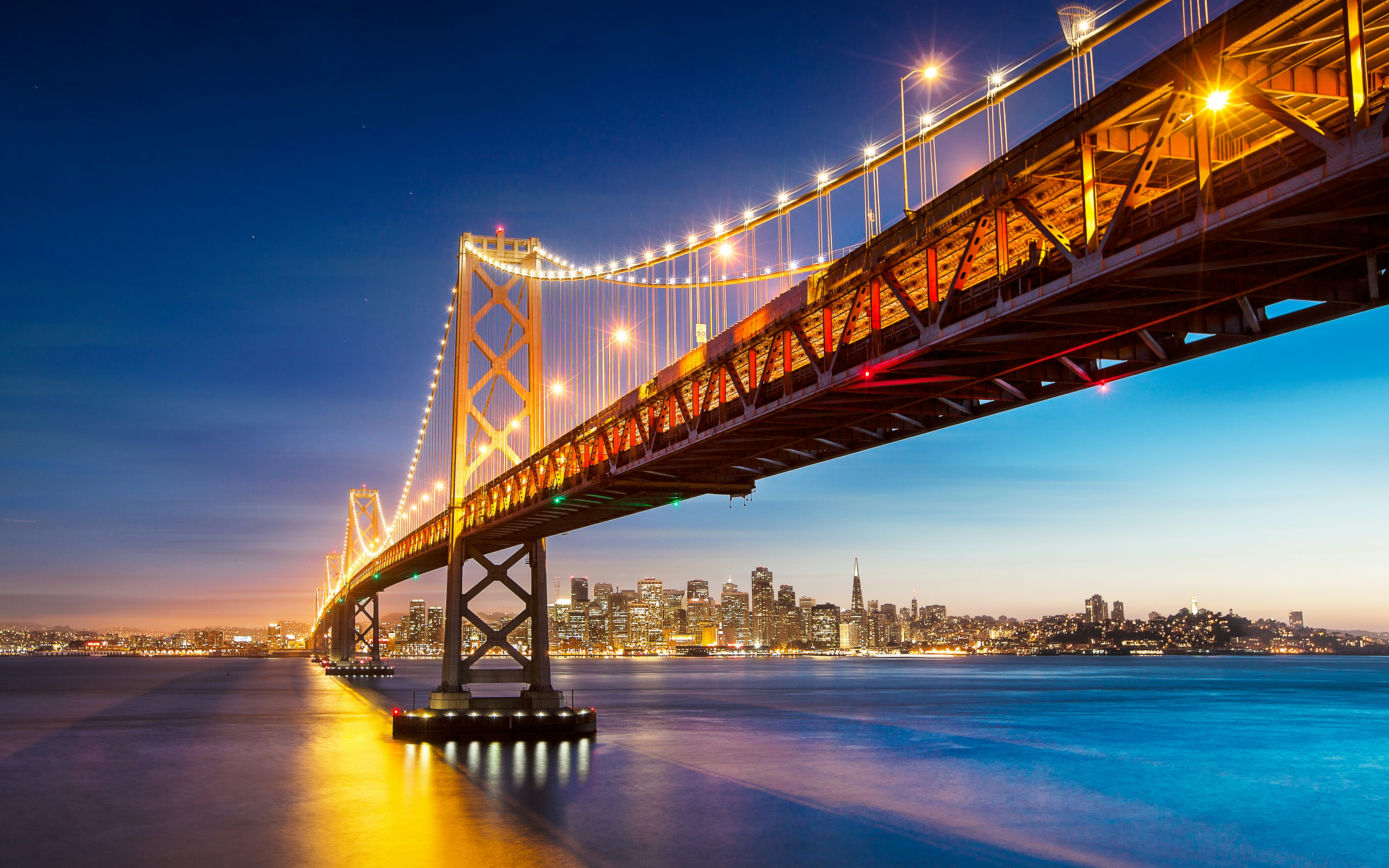 Evening view of a lighted Bay Bridge with the San Francisco skyline in the distance.
