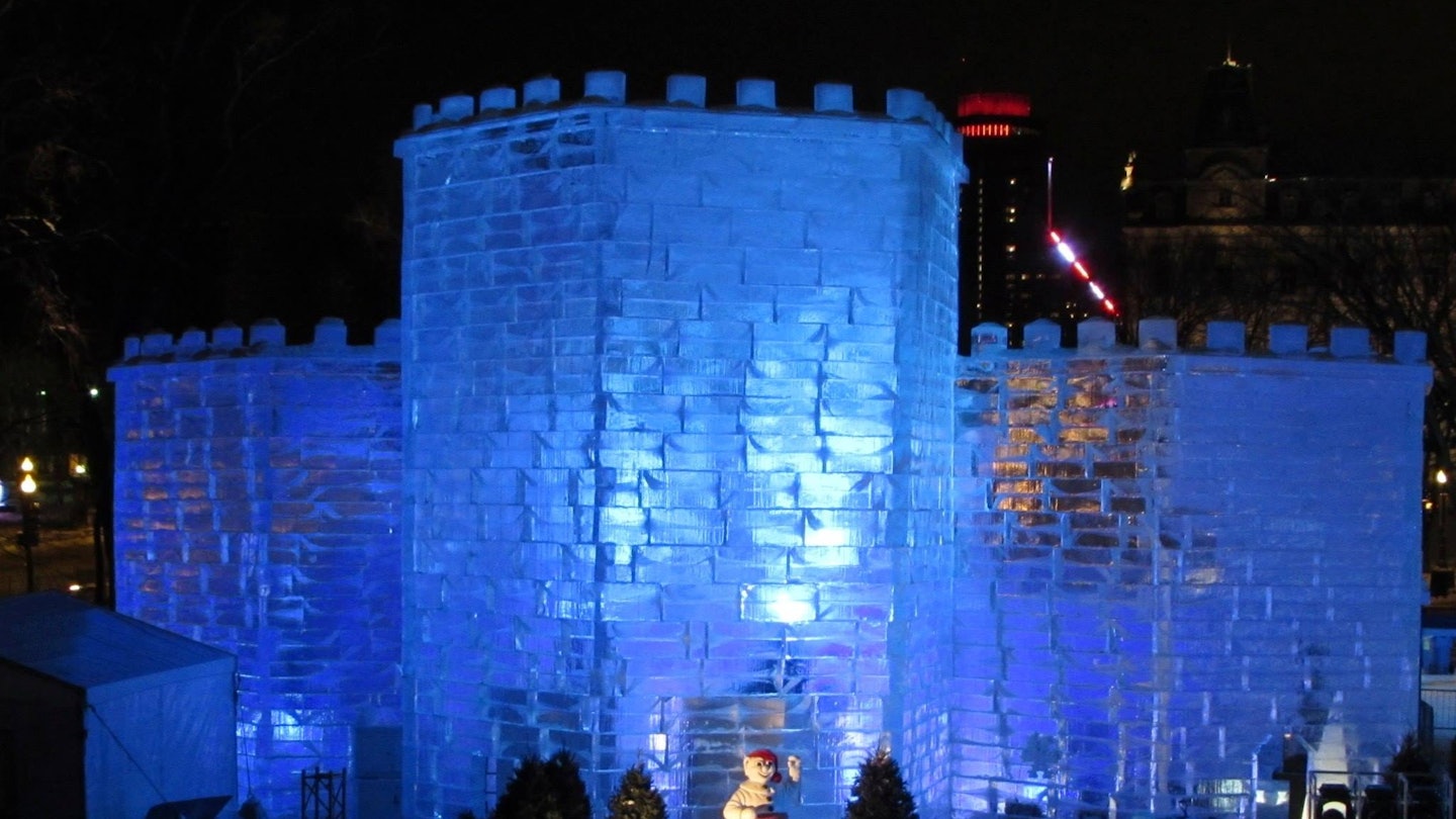 A transparent castle made of giant ice blocks is lit with a blue light from inside, against a pitch black sky.